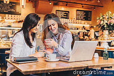 Two young happy women are sitting in cafe at table in front of laptop, using smartphone and laughing. Stock Photo