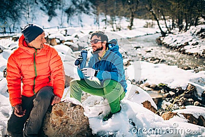 Two young male tourists talk while drinking coffee near a river in the mountains. Stock Photo