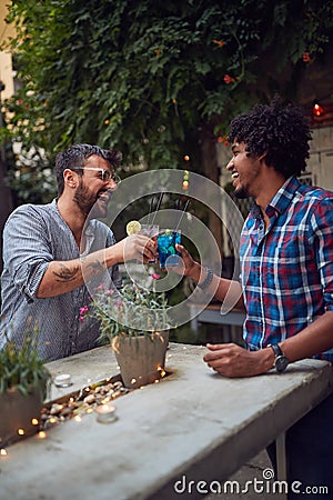 Two young multiethnic male laughing and toast with cocktails in outdoor cafe. multiethnic, friendship, socializing, leisure, Stock Photo
