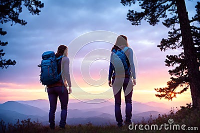Two young non recognizable trekking woman with backpacks standing on top of mountain at sunset and enjoying view. AI Stock Photo