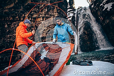 Two young professional male tourists are preparing to camp in the mountains near the river in winter. Stock Photo