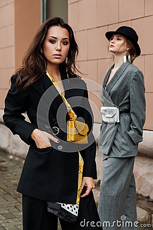 Two young stylish beautiful women fashion models are posing in street, wearing pantsuit, hat, having purse on waist. Stock Photo