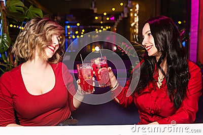 Two young women in red clothes drink cocktails and celebrate in a nightclub or bar. Women clink glasses Stock Photo