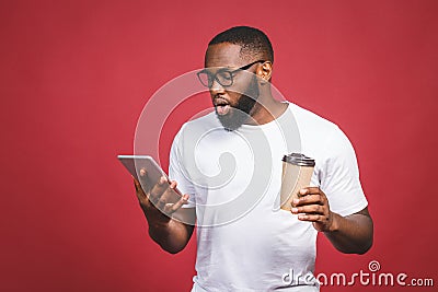 Typing a message. Surprised black man typing something on the mobile phone, drinking coffee while standing isolated against red Stock Photo