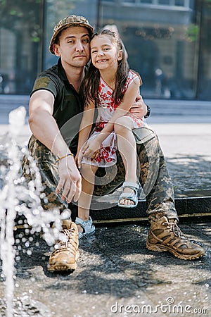 Ukrainian soldier returning from the war meets his happy little daughter and embraces her near fountain in city Stock Photo