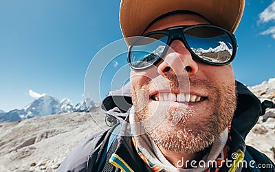 Ultra-wide lens angle portrait shot of high altitude mountain smiling unshaven happy hiker in baseball cap with snow peaks and Stock Photo