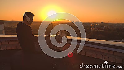 Um programador com um laptop em fones de ouvido senta no telhado e escreve código no pôr do sol e ouve música Trabalho remoto video estoque Um programador com um laptop em fones de ouvido senta no telhado e escreve código no pôr do sol e ouve música Trabalho remoto video estoque