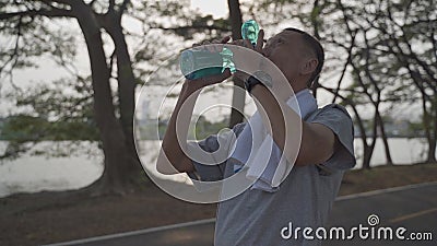 Un homme âgé et assoiffé d'activité asiatique boit de l'eau fraîche à la bouteille dans le parc d'arbres pendant le coucher du sol banque de vidéos Un homme âgé et assoiffé d'activité asiatique boit de l'eau fraîche à la bouteille dans le parc d'arbres pendant le coucher du sol banque de vidéos