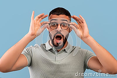 Unbelievable. Shocked middle eastern man touching his glasses and opening mouth, looking at camera on blue background Stock Photo