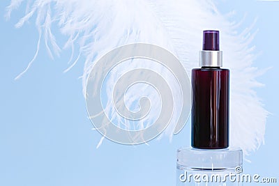 Unbranded brown spray bottle on glass podium and large white ostrich feather on blue background. Sunscreen. Blank label bottle for Stock Photo
