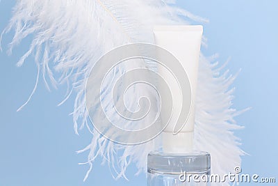 Unbranded white squeeze bottle cream tube on glass podium and large white ostrich feather on blue background. Lotion, balsam, hand Stock Photo