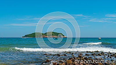 Under the blue sky and white clouds, the waves wash on the beach, and the small island in the distance Stock Photo