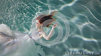 Underwater girl. Beautiful red-haired woman in a white dress, swimming under water. Stock Photo
