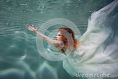 Underwater girl. Beautiful red-haired woman in a white dress, swimming under water. Stock Photo