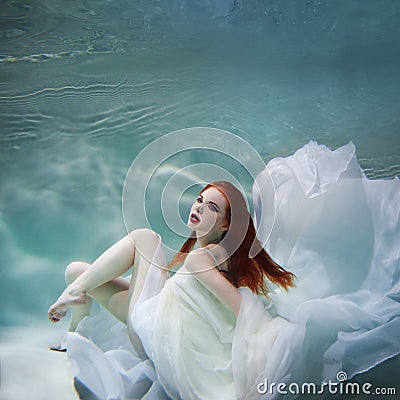 Underwater girl. Beautiful red-haired woman in a white dress, swimming under water. Stock Photo