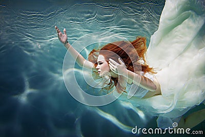 Underwater girl. Beautiful red-haired woman in a white dress, swimming under water. Stock Photo