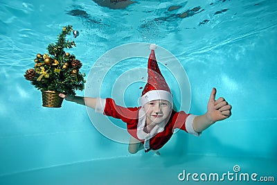 Underwater photo with a Christmas tree of a cheerful little boy in a Santa Claus costume. He dives to the bottom of the Stock Photo