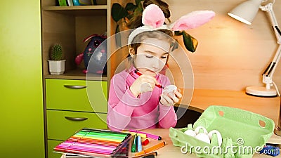 Une fille avec des oreilles de lapin sur la tête peint des oeufs blancs avec des stylos à pointe feutrée à la table. préparation à banque de vidéos Une fille avec des oreilles de lapin sur la tête peint des oeufs blancs avec des stylos à pointe feutrée à la table. préparation à banque de vidéos