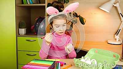 Une fille avec des oreilles de lapin sur la tête peint des oeufs blancs avec des stylos à pointe feutrée à la table. préparation à clips vidéos Une fille avec des oreilles de lapin sur la tête peint des oeufs blancs avec des stylos à pointe feutrée à la table. préparation à clips vidéos