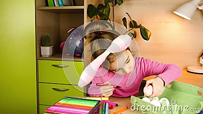 Une fille avec des oreilles de lapin sur la tête peint des oeufs blancs avec des stylos à pointe feutrée à la table. préparation à banque de vidéos Une fille avec des oreilles de lapin sur la tête peint des oeufs blancs avec des stylos à pointe feutrée à la table. préparation à banque de vidéos