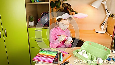 Une fille avec des oreilles de lapin sur la tête peint des oeufs blancs avec des stylos à pointe feutrée à la table. préparation à clips vidéos Une fille avec des oreilles de lapin sur la tête peint des oeufs blancs avec des stylos à pointe feutrée à la table. préparation à clips vidéos