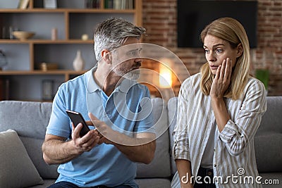 Unhappy adult caucasian man with beard shows phone to shocked lady, woman suffers from gadget addiction Stock Photo