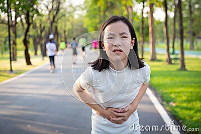 Unhappy asian little cute girl holding her hands on belly,suffers from stomach pain,colic after running,exercise in outdoor park, Stock Photo