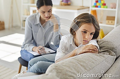 Unhappy child sitting on sofa during therapy session with therapist or psychologist Stock Photo