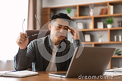 Unhappy mature asian guy analyzes data on laptop, presses hand to head, suffers from problems in business Stock Photo