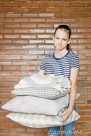 Unhappy preteen girl holding stack of pillows and blankets against brick wal. Homework, cleaning Stock Photo
