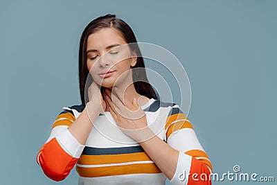 Unhealthy ill girl touches inflamated throat, suffers from pain, has eyes shut, wears casual clothing, stands indoor against blue Stock Photo