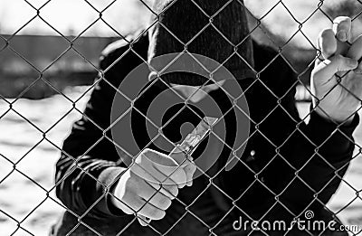 Unidentifiable teenage boy behind wired fence holding a paper knife at correctional institute, focus on the fence in black and whi Stock Photo