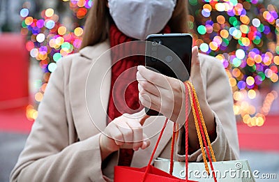 Unidentified woman with protective mask using mobile phone for shopping online and carrying bags on Christmas time. Focus on the Stock Photo