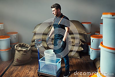 Uniformed worker weighs roasted coffee in a transparent tray on a scale in a coffee industry warehouse with bags and white buckets Stock Photo