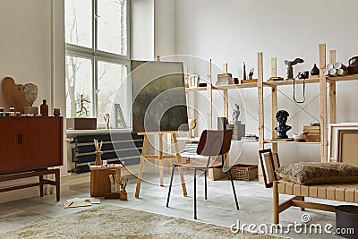 Unique artist workspace interior with stylish teak commode, wooden easel, bookcase, artworks, painting accessories, decoration. Stock Photo