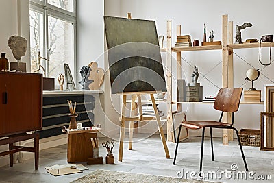 Unique artist workspace interior with stylish teak commode, wooden easel, bookcase, artworks, painting accessories, decoration. Stock Photo