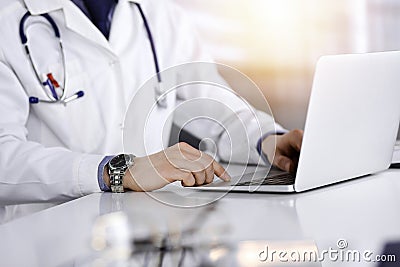 Unknown male doctor sitting and working with laptop in a darkened clinic, glare of light on the background, close-upof Stock Photo