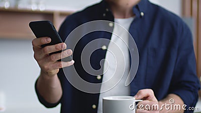 Unknown man typing on smartphone on kitchen. Unrecognizable man drinking tea. Stock Photo