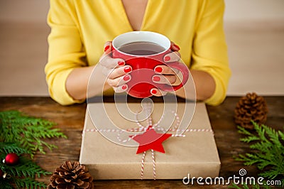 Unrecognisable woman sitting at a table with beautifully wrapped christmas present, holding cup of tea. Gift wrapping concept. Stock Photo