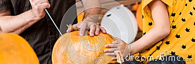 Unrecognisable young girl sitting on kitchen table, helping her father to carve large pumpkin. Halloween family banner. Stock Photo