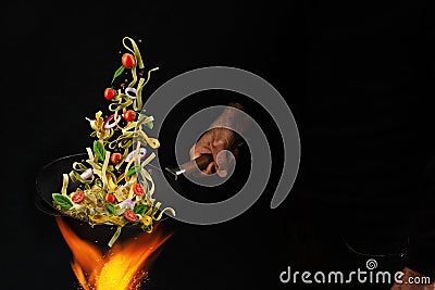 Unrecognizable man holding wok pan above fire and cooking pasta with cherry tomatoes, onion and basil against black Stock Photo