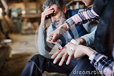 A woman bandaging a hand of a man worker after accident in carpentry workshop. Stock Photo