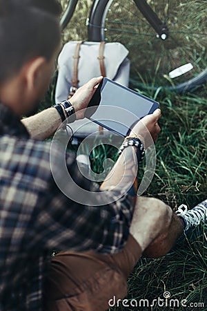 Unrecognizable Young Man Cyclist Sits On Summer Meadow Near Bicycle, Holding And Looking At Tablet Recreation Resting Travel Stock Photo