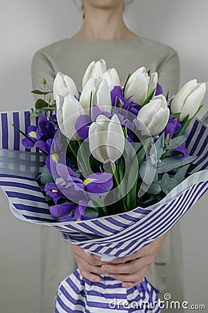 Unrecognizible woman in beige dress holding bouquet of white tulips and violet iris in striped lilac wrapping paper. Stock Photo