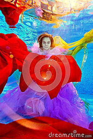 An unusual portrait of a beautiful girl who poses underwater surrounded by colorful fabrics and rays of light Stock Photo