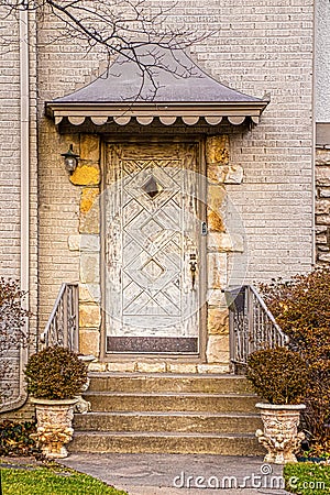 Unusual rustic wooden door surrounded by rock trim in light colored upscale brick house with awning on winter day with all bushes Stock Photo