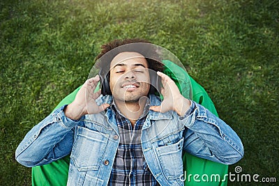 Upper view portrait of pleased relaxed african-american man with bristle lying on grass while listening music with Stock Photo