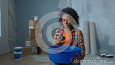 Upset African American woman is holding blue bowl into which drops of water are dripping from ceiling leak. Female is Stock Photo