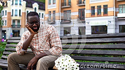 Upset afro-american man sitting lonely on city bench with bouquet, failed date Stock Photo