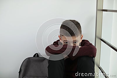 Upset boy with backpack sitting near wall indoors. Stock Photo